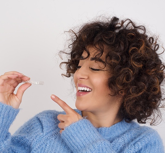 A woman with curly hair and wearing a blue sweater holds an Invisalign aligner in her right hand while smiling