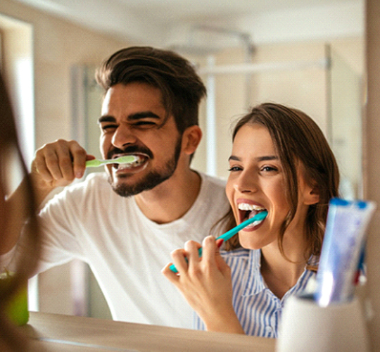 A man and woman brushing their teeth in the mirror as a way to care for their veneers