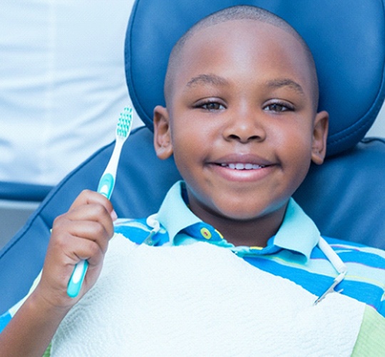 A young boy wearing a striped shirt holds a manual toothbrush and smiles during a regular appointment
