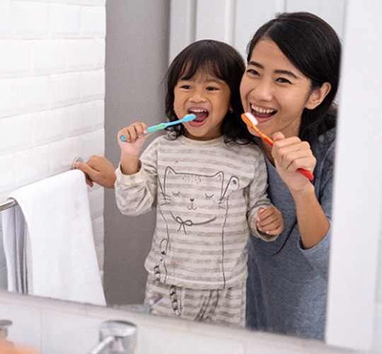 A mother and daughter stand in front of the mirror in their bathroom and brush their teeth