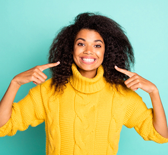 A young woman wearing a yellow sweater and pointing to her new and improved smile