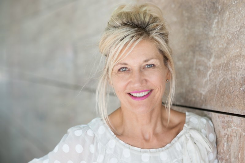 an older woman with her hair up smiling while leaning against a wall after receiving her dental implants